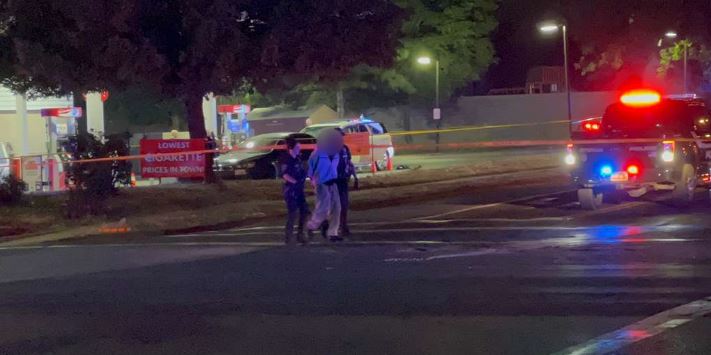 Nighttime street scene with a police car flashing red and blue lights, yellow caution tape, and a group of people near a gas station. Umbrellas suggest an incident scene.