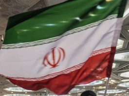 Iranian flag draped indoors at an event, green white red with the emblem in the white band, hanging in a hall.