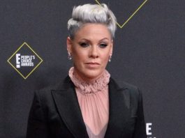 Woman with short silver hair in a black blazer and pink ruffled blouse posing on the red carpet at the People’s Choice Awards.