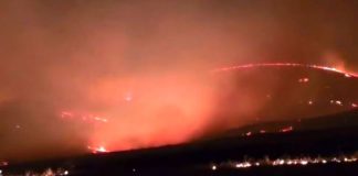 Nighttime wildfire along a ridge with flames and smoke lighting the sky.