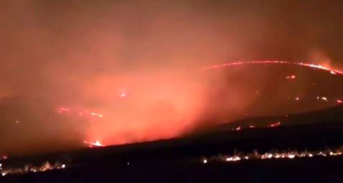 Tooele Nighttime wildfire along a ridge with flames and smoke lighting the sky.