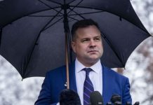 A man in a blue suit stands under a large umbrella behind several microphones, speaking at a press conference outdoors.