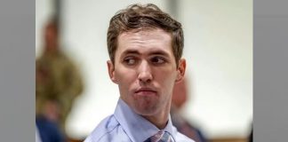 Side profile of a young man in a light blue dress shirt and pink striped tie, looking to his left in a blurred office or conference room setting.