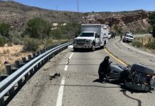 Motorcycle lying on its side in a rural two-lane highway; an ambulance and another vehicle stopped nearby with desert hills in the background.