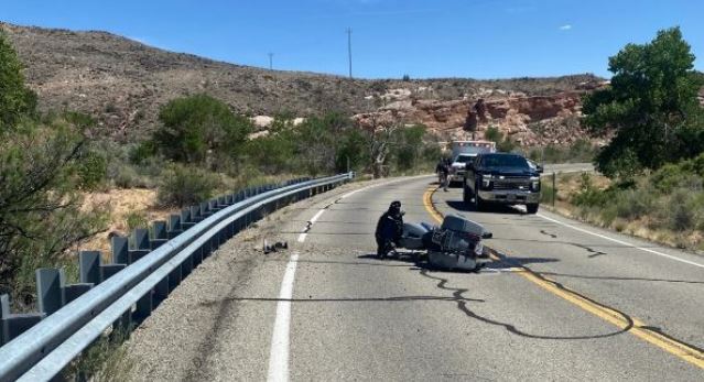 Motorcycle crash on a winding desert road; rider on the pavement next to a damaged bike, a pickup truck stopped nearby, guardrail on left.