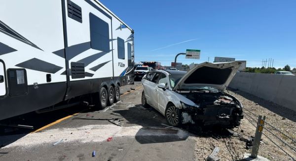 White sedan with severe front-end damage beside a large RV trailer on a highway shoulder.