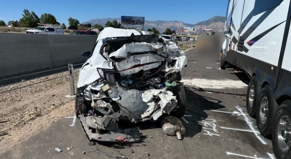 White car with severely crushed front end after a crash on the roadside, debris scattered nearby, with a trailer parked to the right.