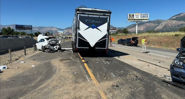Highway crash scene with a large RV trailer stopped in the middle, a severely damaged white car to the left, debris scattered across the road, and responders nearby.