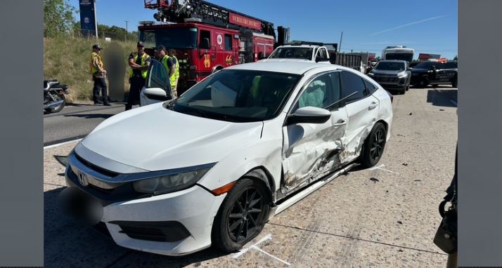 White sedan with significant side damage sits on a highway; emergency responders and a red fire truck are in the background.