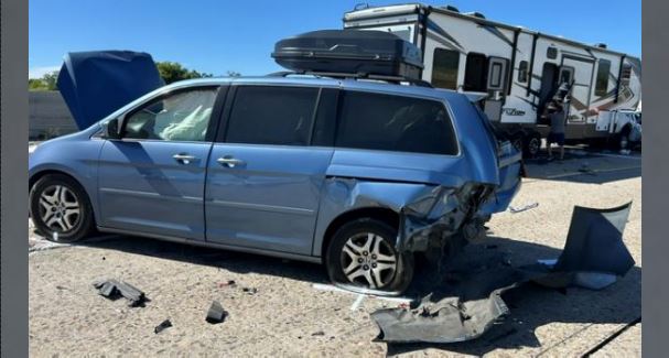 Blue minivan with severe rear-right damage on a gravel lot, debris scattered nearby, with a large RV trailer parked in the background.