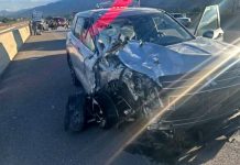 Silver SUV with severe front-end damage on a highway, debris around and responders visible in the distance