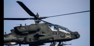 AH-64 Apache attack helicopter in flight, with landing gear visible and weapons mounted under the fuselage.