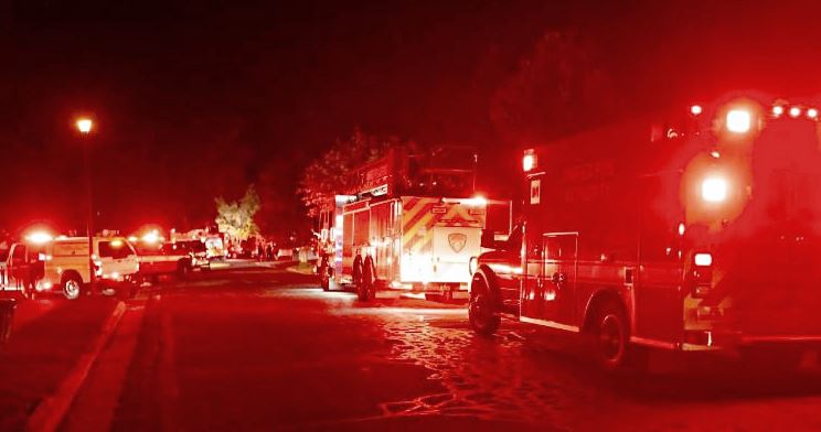 Nighttime street scene with several fire trucks and red emergency lights flashing along the curb.