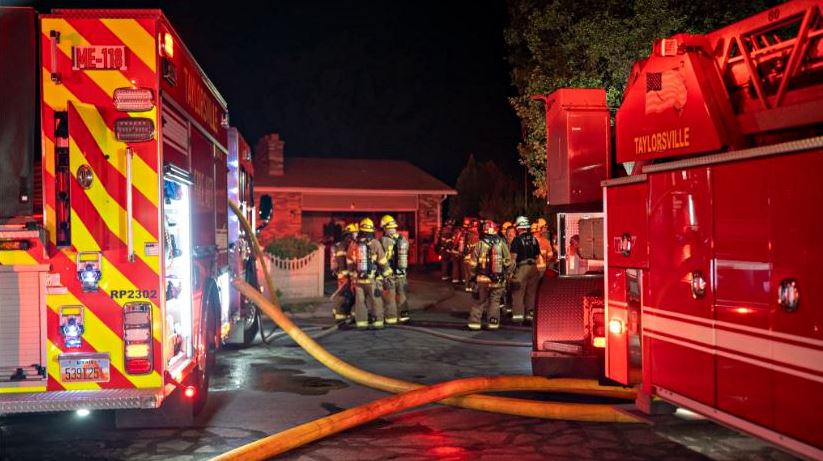 Nighttime scene of firefighters with two red fire engines, laying yellow hoses across a residential street.