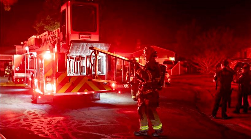 Firefighters in turnout gear stand beside a red fire engine with flashing lights at night on a residential street.