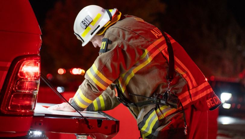 Firefighter in turnout gear and helmet writing on a clipboard at the back of a red fire truck during a nighttime emergency.