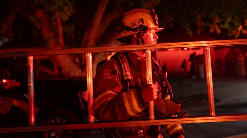Firefighter in turnout gear and helmet carrying a ladder during a nighttime emergency scene, red emergency lighting nearby.