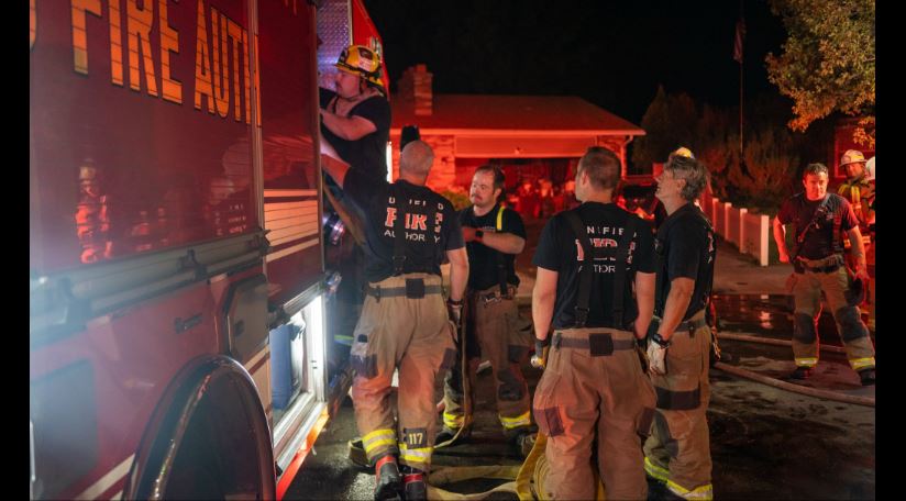 Firefighters in turnout gear stand beside a red fire truck at night, preparing equipment outside a building.