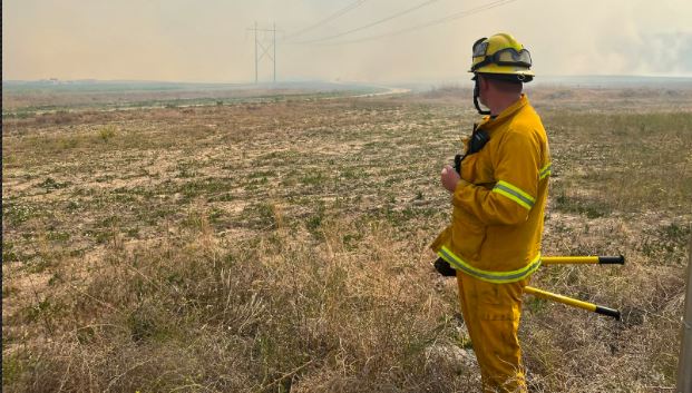 Firefighter in yellow turnout gear stands in a dry grass field, monitoring distant smoke from a grass fire with equipment at his side and power lines in the background.