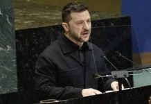 Man in a black jacket speaks at a podium with microphones in a formal assembly hall, behind a clear divider.