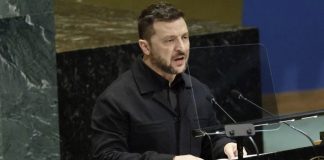 Man in a black jacket speaks at a podium with microphones in a formal assembly hall, behind a clear divider.