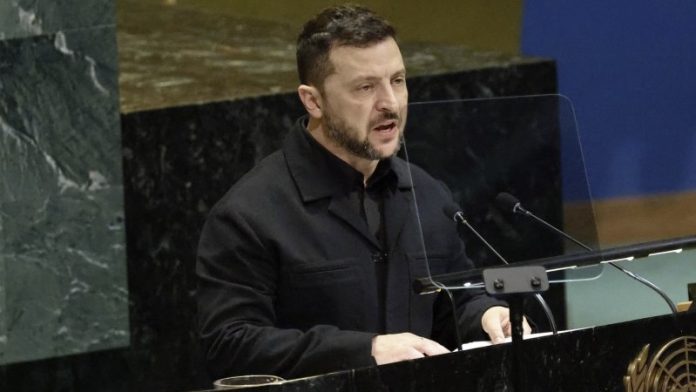 Man in a black jacket speaks at a podium with microphones in a formal assembly hall, behind a clear divider.