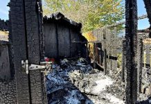 Charred wooden frame of a burned house, ashes on the floor, blue sky visible through damaged walls after a fire reconstruction scene