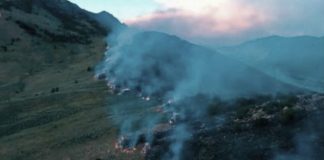 Smoke from a wildfire along a rolling hillside, with blue-tinged smoke drifting over mountains at dusk and small flames visible near the slope edge.