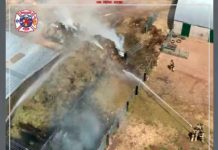 Aerial view of firefighters battling a blaze along a chain-link fence beside a building, with dense smoke and flames visible nearby.
