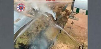 Aerial view of firefighters battling a blaze along a chain-link fence beside a building, with dense smoke and flames visible nearby.
