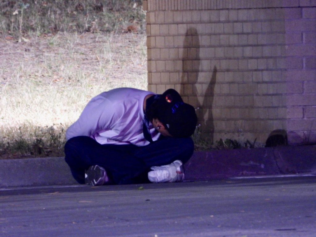 bf4d1dca-e89d-4748-bb0c-34c62c423049 | Gephardt Daily Person crouched on a curb with head bowed against a brick wall at night, wearing a white shirt and dark pants, looking down in distress/solitude.
