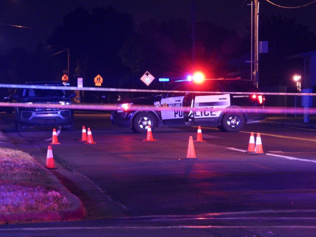 c9a5e787-c08f-4031-aa6d-dfc94833d753 | Gephardt Daily Nighttime police scene with a patrol SUV, orange cones, and police tape across a road; red and blue emergency lights illuminating the area at night.