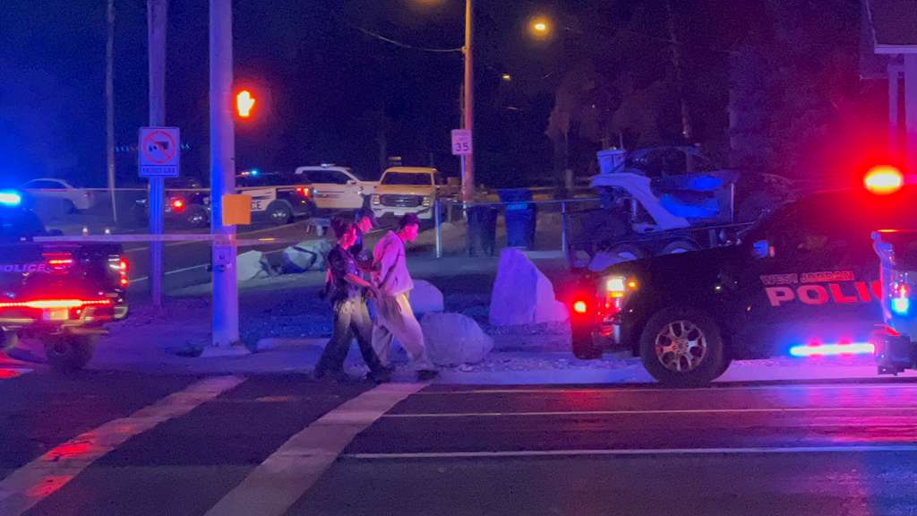 e3aefa9c-2876-41c8-bdcf-29fd5ede10b6 | Gephardt Daily Night scene at an intersection with police cars flashing red and blue lights, yellow tape, and two people being escorted by an officer near a curbside mass of rocks or debris.