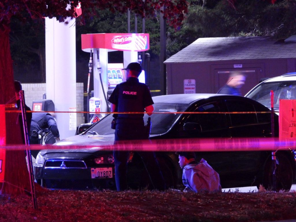 e74b3c8e-ad19-4e7f-9ad6-4c400ebd7230 | Gephardt Daily Police scene at a gas station with a black car and orange tape; officer stands with back to camera studying the area