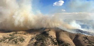 Dry, arid hills with sparse shrubs as a large plume of wildfire smoke drifts across the landscape under a blue sky.
