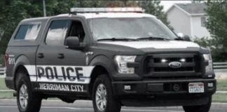 Police pickup truck with 'POLICE' and 'HERRIMAN CITY' markings driving on a street, emergency lights on the roof.