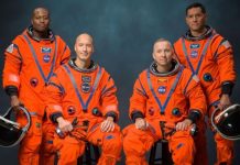 Group of four astronauts in bright orange spacesuits posing for a studio portrait, two standing at the ends and two seated, helmets in hand or by their sides.