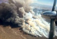 Aerial view from a propeller plane showing a large wildfire with dark smoke and flames rising over a desert landscape below.