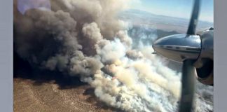 Aerial view from a propeller plane showing a large wildfire with dark smoke and flames rising over a desert landscape below.