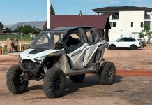 White and gray Polaris RZR off-road side-by-side parked on a dirt lot, muddy and ready for a ride, with buildings and a white SUV in the background.