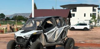 White and gray Polaris RZR off-road side-by-side parked on a dirt lot, muddy and ready for a ride, with buildings and a white SUV in the background.