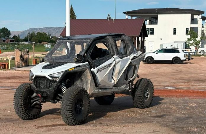 White and gray Polaris RZR off-road side-by-side parked on a dirt lot, muddy and ready for a ride, with buildings and a white SUV in the background.