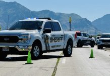 White sheriff pickup with blue emergency lights leading a convoy of police vehicles on a mountain highway, with green cones in the road.