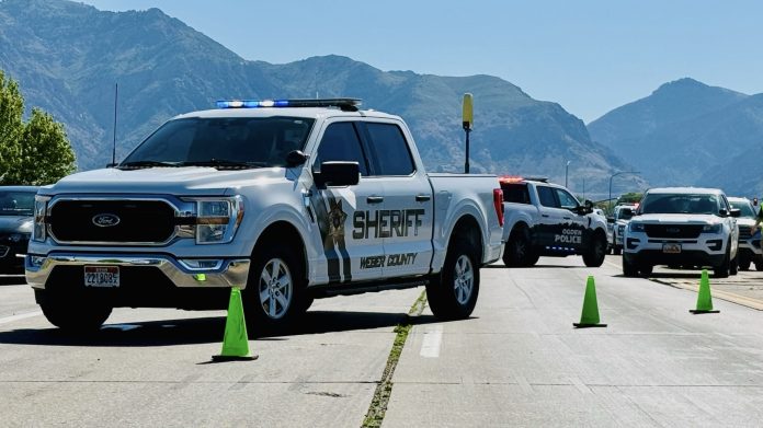 White sheriff pickup with blue emergency lights leading a convoy of police vehicles on a mountain highway, with green cones in the road.
