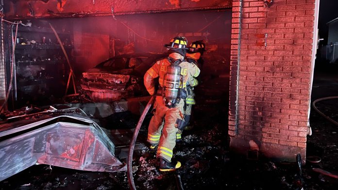 Two firefighters in full turnout gear stand inside a charred garage, inspecting a burned car amid red glow and smoke.