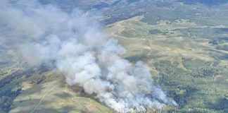 A wildfire burns on grassy hillside with thick smoke drifting over the landscape from above.