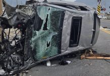 Upside-down silver SUV with a shattered windshield and exposed engine after a crash, debris scattered on the road at an intersection.