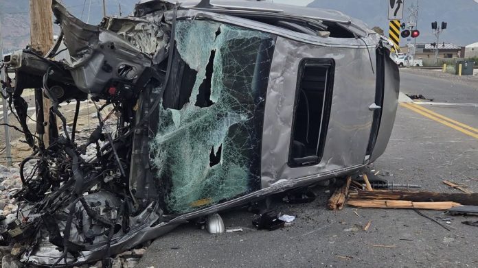 Upside-down silver SUV with a shattered windshield and exposed engine after a crash, debris scattered on the road at an intersection.