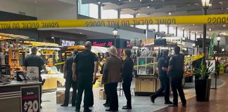 Police officers stand inside a taped-off area at a shopping center food court, inspecting stalls behind bright yellow caution tape.