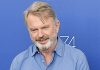 Actor Sam Neill of ‘Jurassic Park’ fame dies at age 78 Smiling older man with gray beard in a light blue button-down shirt against a blue backdrop.